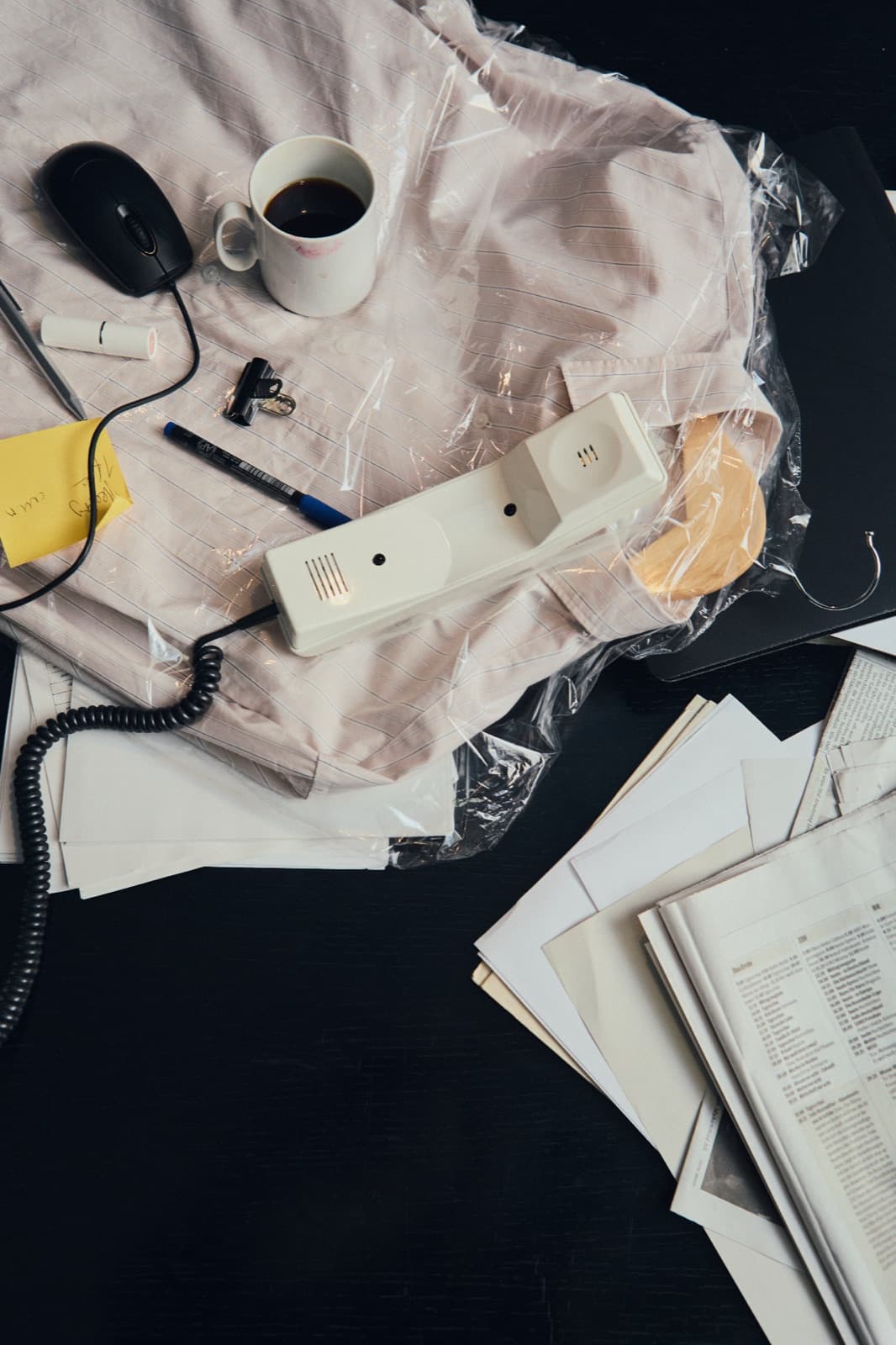 Mesa bagunçada vista de cima: fone de telefone com fio, caneca de café e mouse sobre uma camisa embalada em plástico, junto a papéis soltos