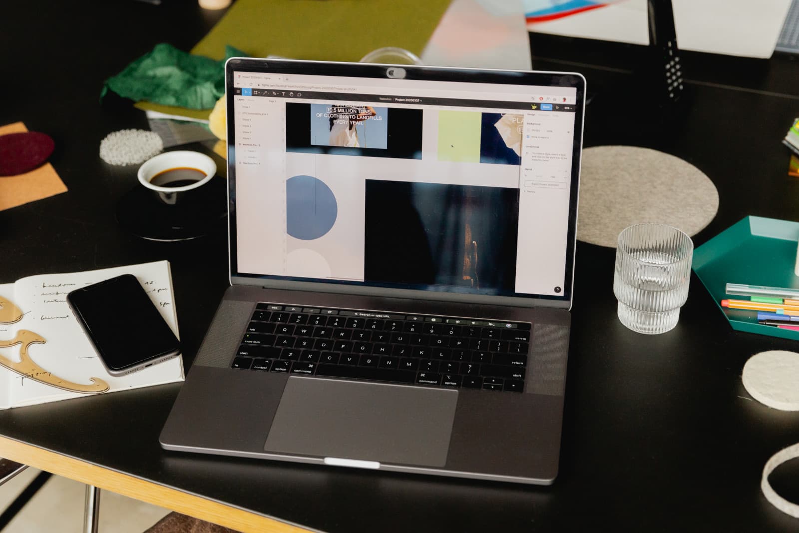 Notebook aberto mostrando um app de design com miniaturas de imagens, junto a caderno de notas à mão, celular, xícara de café e copo d'água